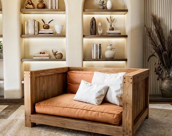May include: A large, wooden armchair with a brown cushion and two white pillows. The armchair is in a room with a light brown rug and white walls. There are shelves with books and decorative objects on the walls.