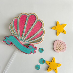 May include: A pink and gold glitter seashell with a turquoise ribbon, two yellow starfish, and a pink seashell with gold glitter accents.