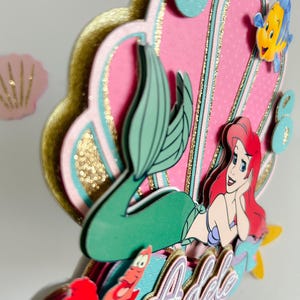 May include: A pink and gold mermaid cake topper with a mermaid, a seashell, and a crab. The mermaid is wearing a green tail and has red hair. The seashell is decorated with gold glitter. The cake topper is personalized with the name "Adele".