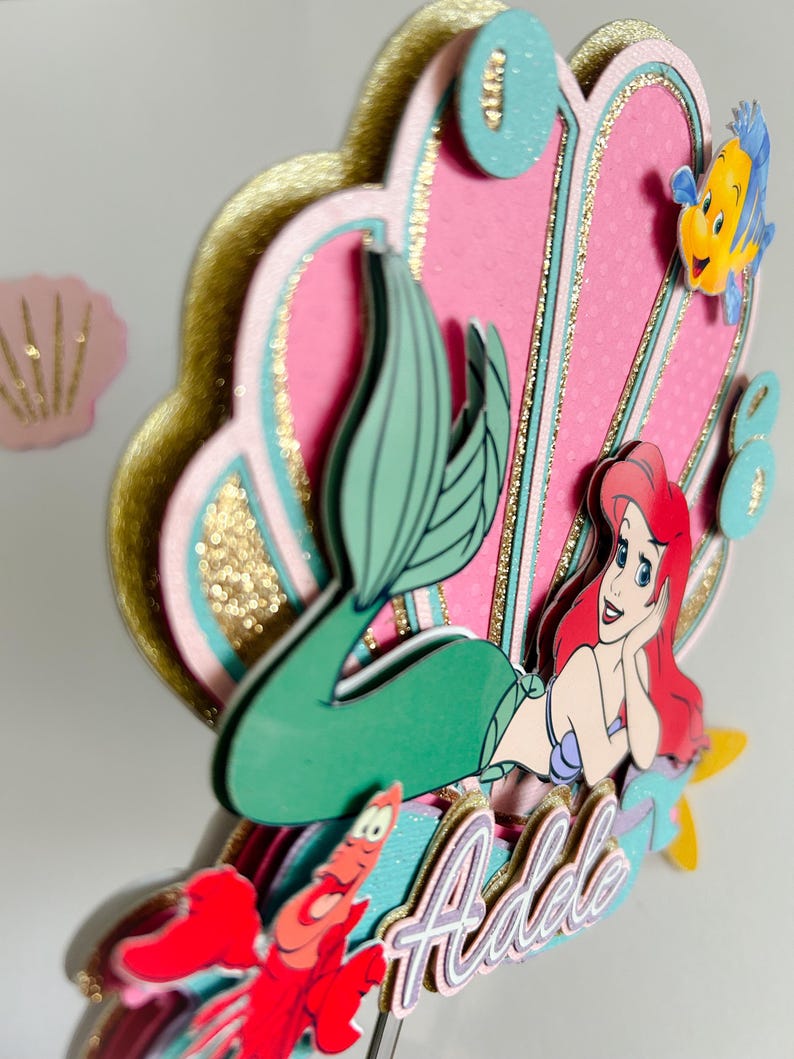 May include: A pink and gold mermaid cake topper with a mermaid, a seashell, and a crab. The mermaid is wearing a green tail and has red hair. The seashell is decorated with gold glitter. The cake topper is personalized with the name "Adele".