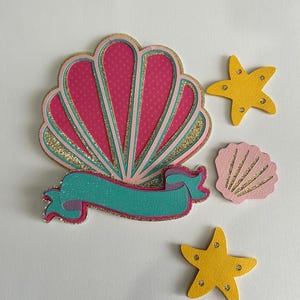 May include: A pink and gold glitter seashell with a turquoise ribbon and two yellow glitter starfish.