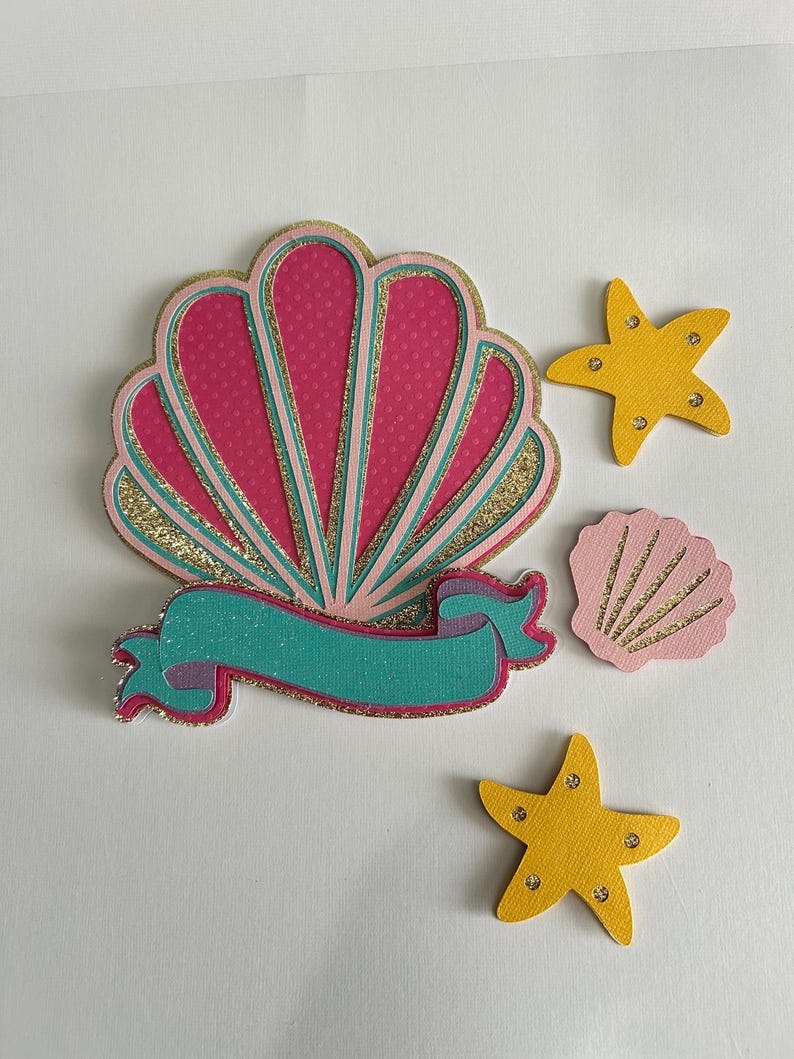 May include: A pink and gold glitter seashell with a turquoise ribbon and two yellow glitter starfish.