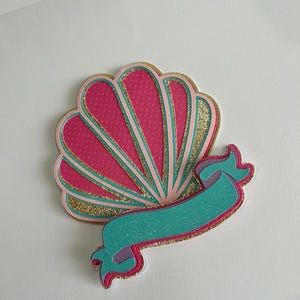 May include: A pink and gold seashell with a teal and purple ribbon. The seashell has a dotted pattern and a glittery finish.