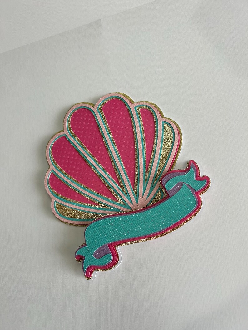 May include: A pink and gold seashell with a teal and purple ribbon. The seashell has a dotted pattern and a glittery finish.