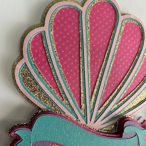 May include: A pink and gold seashell with a teal ribbon. The seashell has a textured surface and is layered with glitter. The ribbon is also layered and has a textured surface.