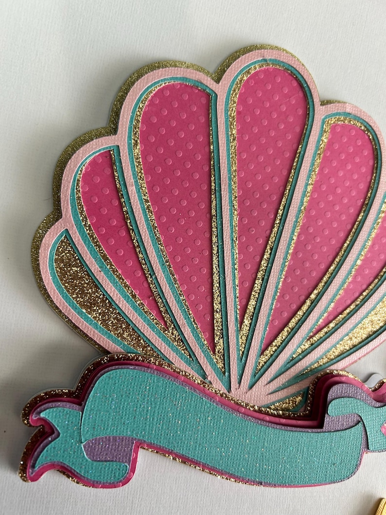 May include: A pink and gold seashell with a teal ribbon. The seashell has a textured surface and is layered with glitter. The ribbon is also layered and has a textured surface.