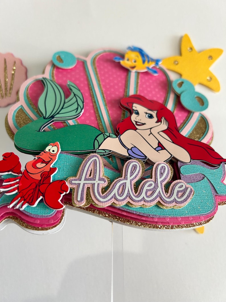 May include: A mermaid-themed cake topper featuring Ariel from The Little Mermaid, a seashell, a crab, a starfish, and a fish. The topper is decorated with glitter and the name "Adele" is written in gold letters.