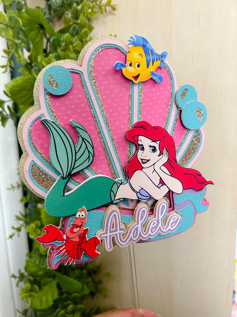 May include: A pink and gold seashell cake topper with a mermaid, a crab, and a fish. The mermaid is wearing a purple seashell bra and has red hair. The cake topper says "Adele" in pink and purple letters.