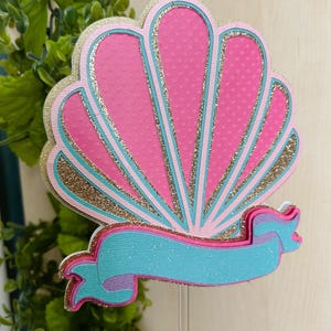 May include: A pink and gold glitter seashell with a turquoise ribbon. The seashell is a cake topper.