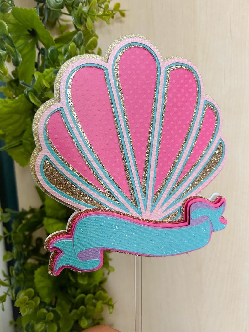 May include: A pink and gold glitter seashell with a turquoise ribbon. The seashell is a cake topper.