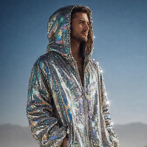 Puede incluir: Chaqueta plateada con capucha y lentejuelas con cierre de cremallera. La chaqueta tiene mangas largas y una capucha. La chaqueta se usa con pantalones oscuros. El fondo es un paisaje desértico.