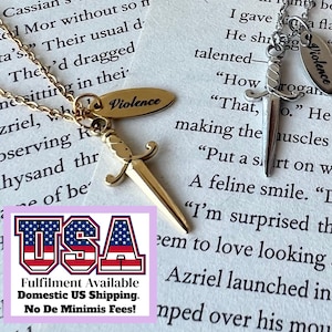 May include: Two necklaces with dagger pendants and the word "Violence" on a small tag. One necklace is gold-toned, the other silver-toned. The pendants are detailed with a sword design. The background is a book page.