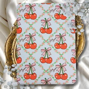 Könnte beinhalten: Hellblauer Buchumschlag mit einem sich wiederholenden Muster aus roten Kirschen mit rosa Schleifen und grünen Stielen, umrahmt von einem floralen Design. Der Text "Book Girlie" wiederholt sich. Der Umschlag liegt auf einem goldfarbenen Tablett.