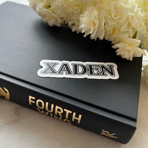 May include: A black and white sticker with the name "Xaden" on a black book with gold lettering that reads "FOURTH WING".