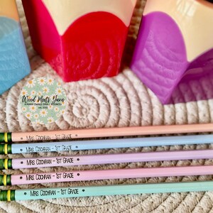 May include: Five colorful pencils with green and black stripes and the text "MRS COCHRAN - 1ST GRADE" printed on each. The pencils are arranged in a row on a woven surface.