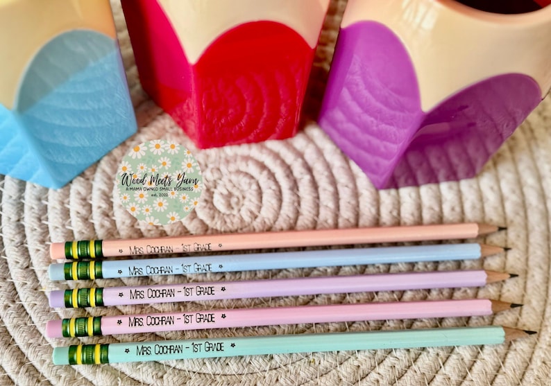 May include: Five colorful pencils with green and black stripes and the text "MRS COCHRAN - 1ST GRADE" printed on each. The pencils are arranged in a row on a woven surface.