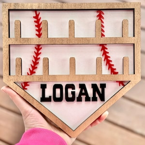 May include: Wooden baseball home plate shaped sign with a white background and red stitching. The sign has eight slots for displaying baseballs and the name "LOGAN" is printed in black letters.