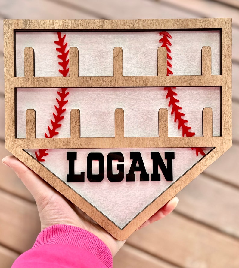 May include: Wooden baseball home plate shaped sign with a white background and red stitching. The sign has eight slots for displaying baseballs and the name "LOGAN" is printed in black letters.