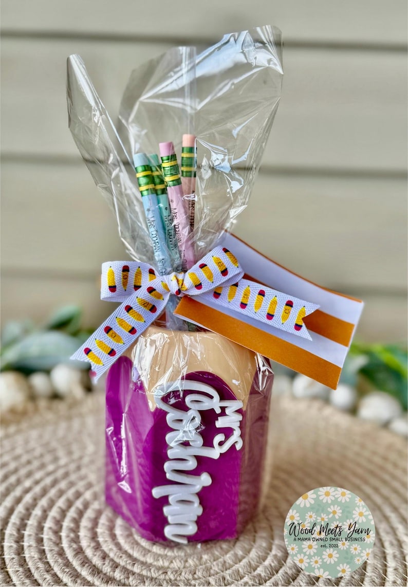 May include: A purple pencil holder with the words "Mrs. Goodman" in white letters. The holder is wrapped in clear cellophane and tied with a yellow and white ribbon. There are several pencils inside the holder.