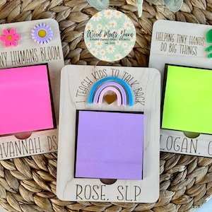 May include: Three wooden note holders with colorful accents and personalized names. The top of each holder features a different design: a flower, a rainbow, and a dinosaur. Each holder has a space for a sticky note.