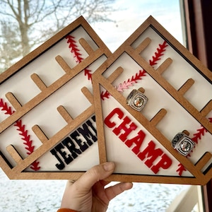 May include: Two wooden baseball-themed ring holders. The holder on the left says "JEREMY" and the holder on the right says "CHAMP". Both holders have red and white baseball stitching and slots for rings.