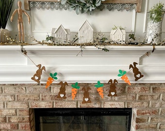 May include: A brown bunny garland with green carrot tops and orange carrots hanging on a white mantel above a brick fireplace.