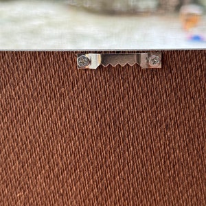 May include: A close-up of a brown canvas textured surface with a silver metal hanging hook attached with two screws.