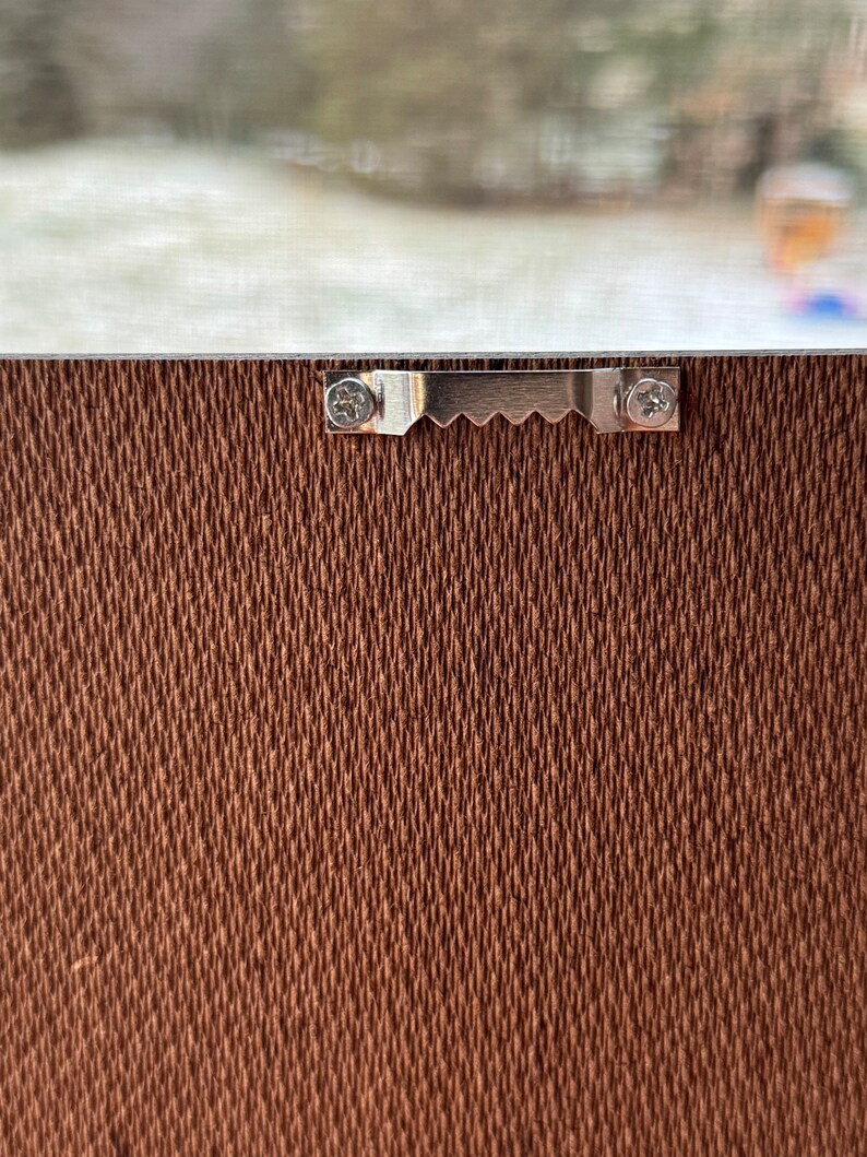 May include: A close-up of a brown canvas textured surface with a silver metal hanging hook attached with two screws.