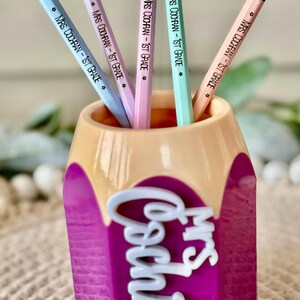 May include: A purple pencil holder shaped like a pencil with the words "Mrs. Cochran" in white letters. The holder has five pencils inside, each with a light blue, pink, or green body and the text "Mrs. Cochran - 1st Grade" written on them.