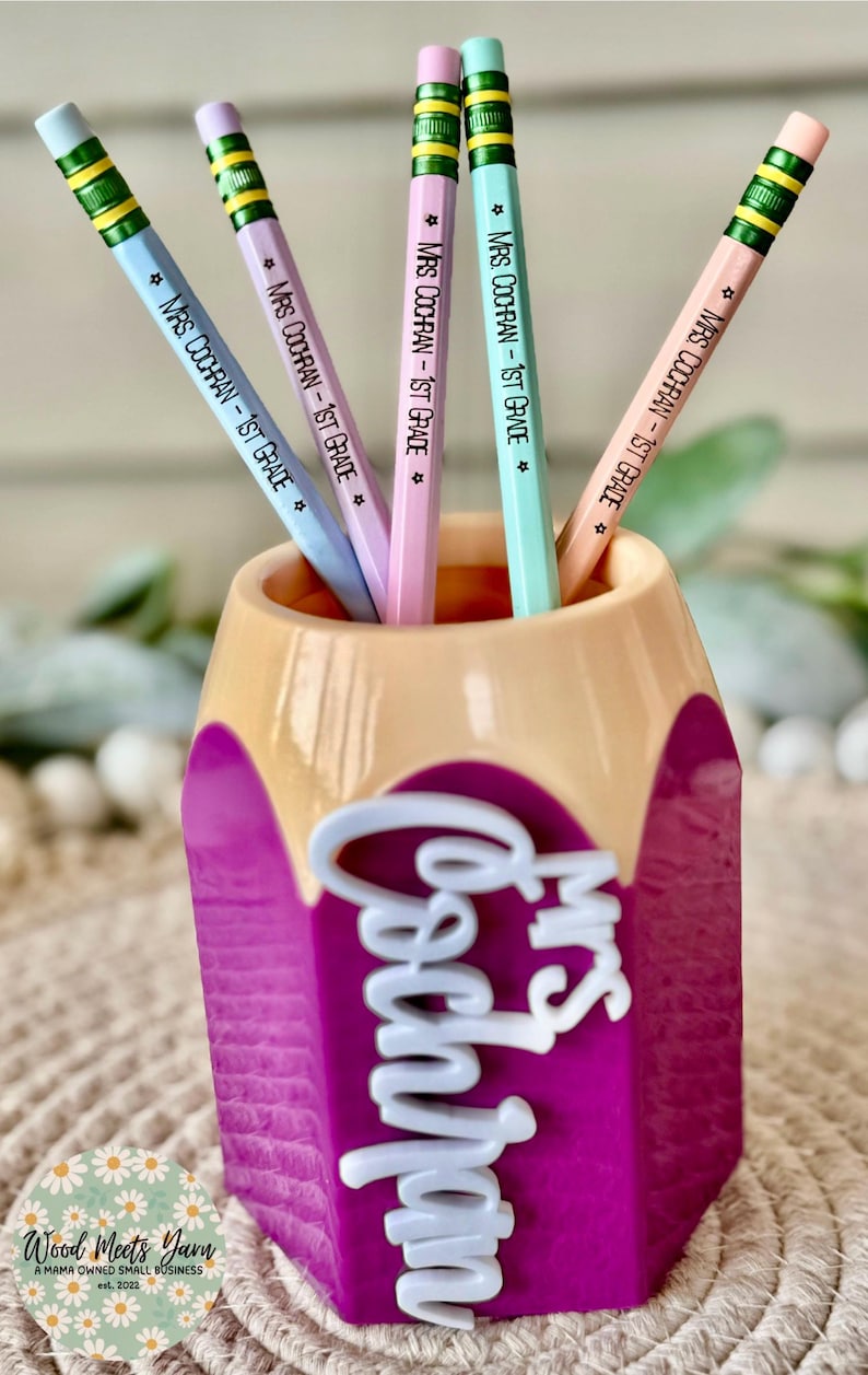May include: A purple pencil holder shaped like a pencil with the words "Mrs. Cochran" in white letters. The holder has five pencils inside, each with a light blue, pink, or green body and the text "Mrs. Cochran - 1st Grade" written on them.