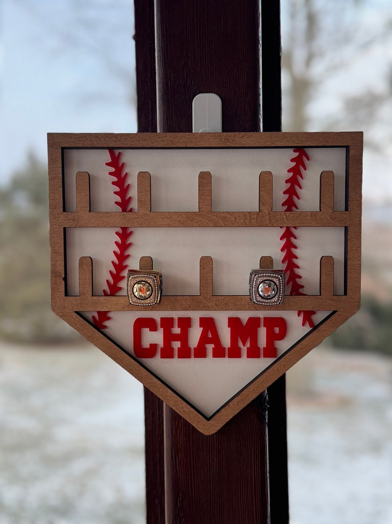 May include: Wooden baseball-themed ring holder with red stitching and the word "CHAMP" in red letters. Two rings are displayed in the holder.