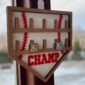 May include: A wooden baseball-shaped ring holder with the word "CHAMP" in red letters. The holder has slots for rings and a baseball stitching design. Two rings are displayed in the holder.