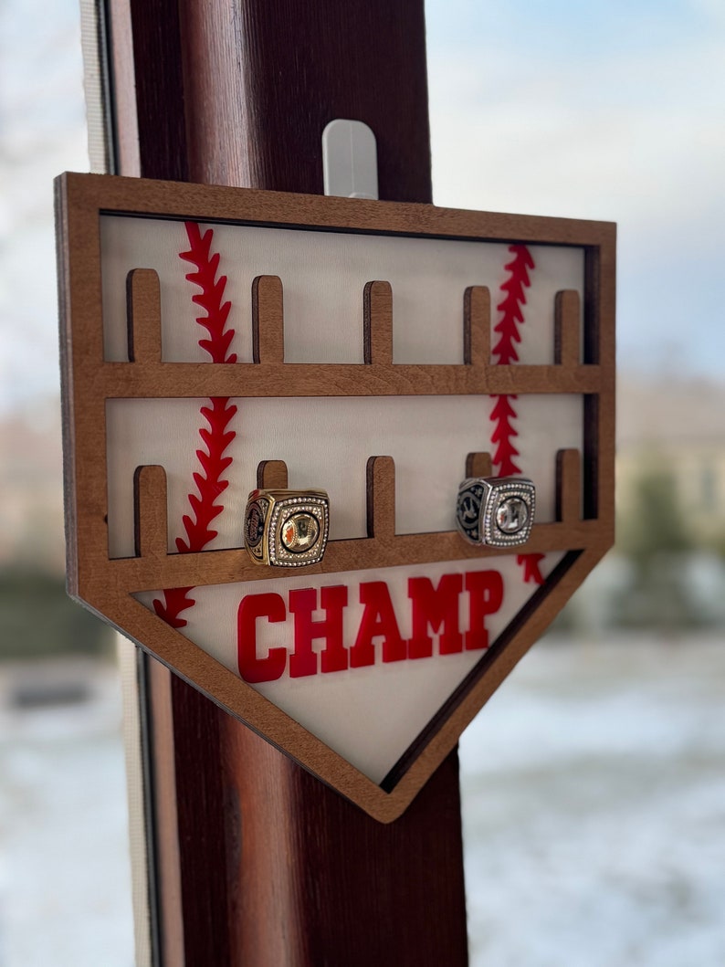 May include: A wooden baseball-shaped ring holder with the word "CHAMP" in red letters. The holder has slots for rings and a baseball stitching design. Two rings are displayed in the holder.