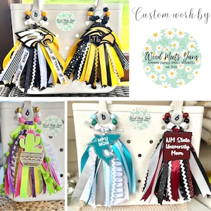 May include: A collection of custom-made bag charms with colorful ribbons and school-themed embellishments. Each charm features a letter "b" and decorative beads. The charms are adorned with school names and mascots, perfect for showing school spirit.