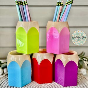 May include: Five colorful pencil-shaped desk organizers in green, pink, blue, red, and purple. Each organizer has a cream-colored top that resembles the eraser of a pencil. The organizers are filled with colorful pencils.