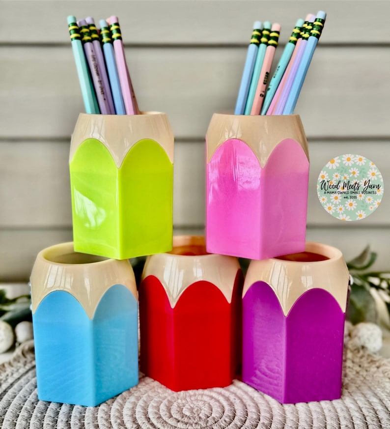 May include: Five colorful pencil-shaped desk organizers in green, pink, blue, red, and purple. Each organizer has a cream-colored top that resembles the eraser of a pencil. The organizers are filled with colorful pencils.