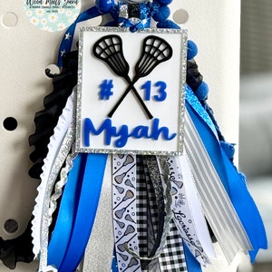 May include: A custom lacrosse mum with a silver glitter frame, crossed lacrosse sticks, and the name "Myah" in blue. The mum features blue, white, silver, and black ribbons, along with a bead accent and the number 13.