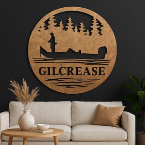 May include: A circular wooden wall sign featuring a laser-cut silhouette of a person fishing from a boat on water, set against a backdrop of pine trees. The name GILCREASE is prominently displayed in bold, black capital letters.