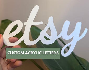 May include: White acrylic "etsy" sign with a cursive font, held in front of a green plant. The sign is in focus, with the text "CUSTOM ACRYLIC LETTERS" in a green rectangle below.