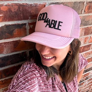 May include: A pink trucker hat with the words "GOD IS ABLE" in black lettering. The hat has a mesh back and a curved brim. The hat is a light pink color.