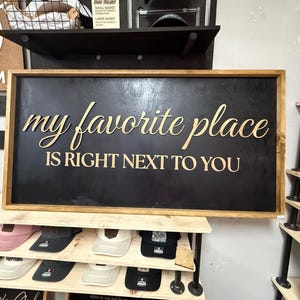 May include: A rectangular wooden sign with a black background and a natural wood frame. The sign features the gold-colored text "my favorite place" in a cursive script above the text "IS RIGHT NEXT TO YOU" in a bold, capitalized sans-serif font.