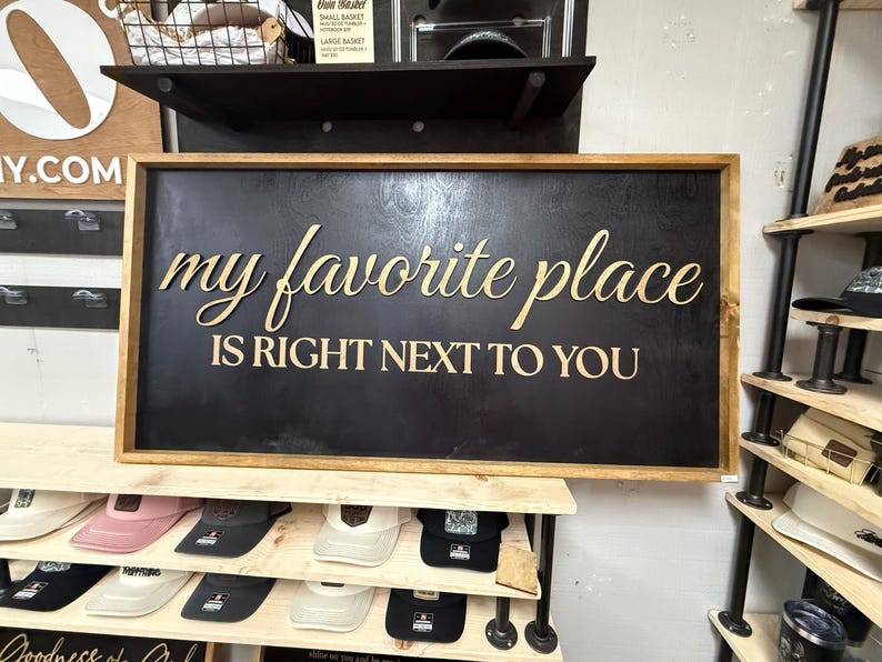 May include: A rectangular wooden sign with a black background and a natural wood frame. The sign features the gold-colored text "my favorite place" in a cursive script above the text "IS RIGHT NEXT TO YOU" in a bold, capitalized sans-serif font.