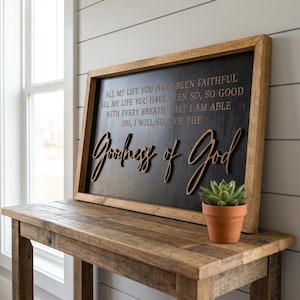 May include: A wooden framed sign with the words "Goodness of God" in a cursive font. The sign is black with the words in a lighter color. The sign is on a wooden table with a small potted plant.