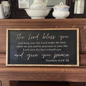 May include: A rectangular wooden sign with a black background and a natural wood frame. The sign features raised, light-colored lettering that reads: The Lord bless you and keep you; the Lord make his face shine on you and be gracious to you; the Lord turn his face toward you and give you peace. Numbers 6:24-26.