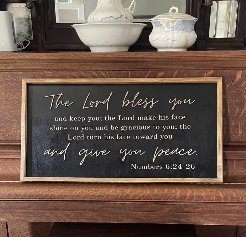 May include: A rectangular wooden sign with a black background and a natural wood frame. The sign features raised, light-colored lettering that reads: The Lord bless you and keep you; the Lord make his face shine on you and be gracious to you; the Lord turn his face toward you and give you peace. Numbers 6:24-26.