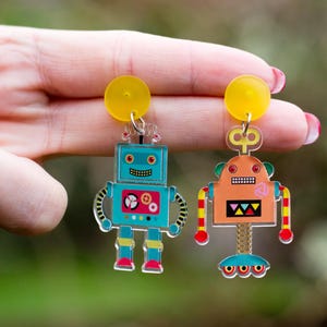 May include: Robot-shaped earrings in teal and orange. The teal robot has red and yellow details, the orange robot has yellow and red details. Both have yellow circular tops and are suspended by silver hooks.