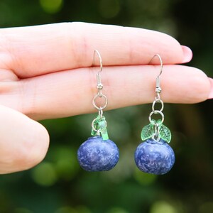 Könnte beinhalten: Ein Paar handgefertigte Ohrringe mit einem Blaubeer-Design. Jeder Ohrring hat einen dunkelblauen, strukturierten Blaubeer-Anhänger mit grünen Blattakzenten und silberfarbenen Ohrhaken. Die Ohrringe werden von einer Hand gehalten.