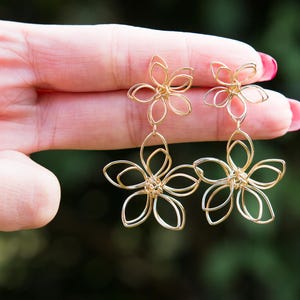 May include: Gold-colored wire flower earrings. Each earring features two flower shapes, one above the other, with delicate wire petals. The earrings are held up against a blurred green background.