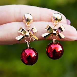 May include: A pair of festive earrings featuring a gold-colored bow and a dangling, shiny red sphere. The earrings have a gold-colored stud at the top. The background is a blurred green.