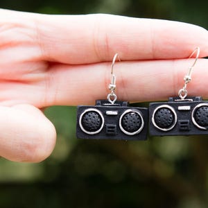 以下が含まれることがあります： 銀色のアクセントが付いた、黒いラジオ型のイヤリングのペア。 イヤリングはレトロなデザインで、スピーカーの詳細と耳用のシルバーフックが特徴です。 赤いマニキュアを塗った手で持っています。